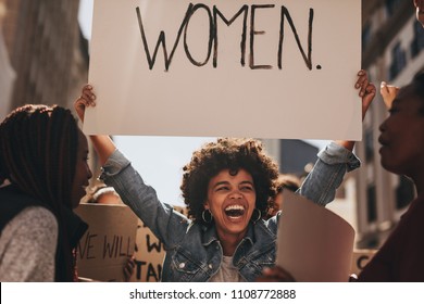 Laughing Young Woman Holding A Banner During A Protest. Group Of Females Activist Protesting On Road For Women Empowerment.