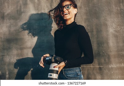 Laughing Young Woman Holding A Dslr Camera Against Brown Background. Successful Female Photographer On A Photo Shoot.