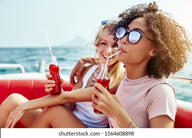 Laughing young woman sipping on a drink while relaxing with her friend on a boat on the ocean during their summer vacation