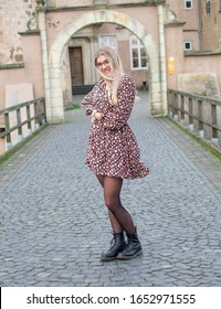 Laughing, young woman wearing glasses with flowing, long, blond hair in a flower dress with laced ankle boots on an old stone bridge