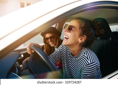 Laughing young woman wearing sunglasses driving a car with her girl friend , close up profile view through the open window