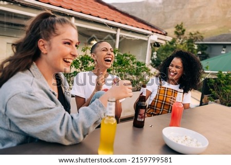 Laughter over cold drinks. Three happy female friends laughing cheerfully while hanging out together on a rooftop. Group of young girlfriends having a good time together on the weekend.