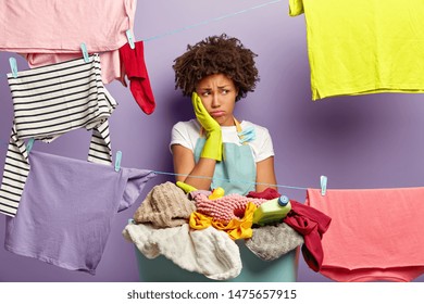 Laundry Day Concept. Fatigue Dissatisfied Woman Touches Cheek And Looks Away In Unhappiness, Stands Near Basket With Pile Of Laundry And Detergent, Washing Lines With Hanged Wet Clothes For Drying