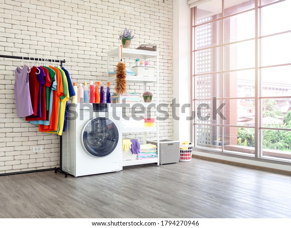 Laundry room interior. Utility room with washing
machine, cleaning equipment, home cleaners, clean wipes, hanging
colorful shirts on clothesline on white brick wall background near
large glass window.
