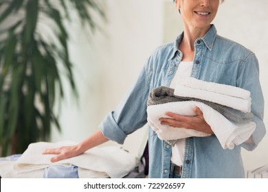 Laundry Service Concept, Close Up Photo Of Woman Holding A Pile Of Ironed Clean Towels Standing At Ironing Board And Smiling 