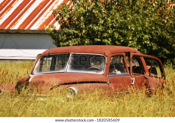 Laurel, DE/USA-September 11, 2020:  Rusted\
out, banged up 80 year old vintage classic car from the late\
1940\'s\'s sits abandoned in an overgrown field with a rusted chicken\
coop roof in the\
background.