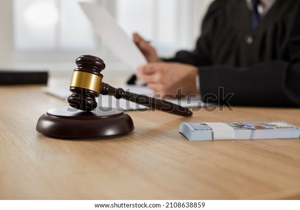 Law, auctioning, corruption, bankruptcy, bail,\
bribery or fines. Close up of judge gavel standing on sound block\
and bundle of dollars on table against background of judge or\
auctioneer with documents