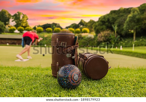 Lawn bowls leather bags and balls on\
the field side while players are playing at\
sunset