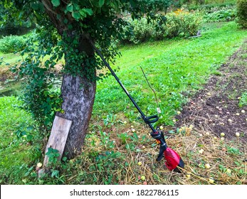 A Lawn Mower Stands Near A Tree. A Mowing Machine Stands Near The Apple Tree, Waiting For The Gardener. Harvesting Yellowed Autumn Grass. Preparing The Garden For Winter.