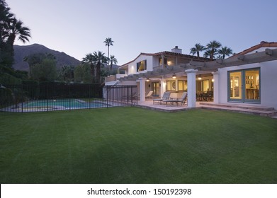 Lawn and swimming pool with lit exterior of home against the sky