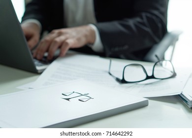 Lawyer Working In Office. Attorney Writing A Legal Document With Laptop Computer. Glasses On Table. Pile Of Paper With Scale And Justice Symbol. Law Firm And Business Concept.