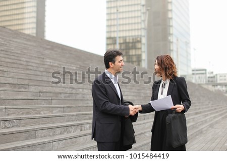 Layer and client standing on big gray stairs. Woman hold documents about court statement in hand. Man tell about very good end of litigation, and bouth smiling look really happy. Concept of pos