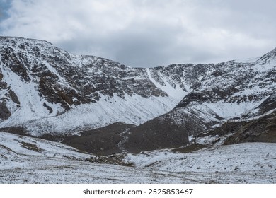 Layered cold landscape with snow-covered grassy, rocky and stony hills against large snowy mountain range under gray cloudy sky. Freshly fallen snow on grass in high mountains in changeable weather. - Powered by Shutterstock