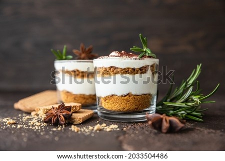 Layered dessert in glass jar with cookie crumble and whipped cream decorated with rosemary and anise, dark brown background. No bake cheesecake, trifle or pudding.