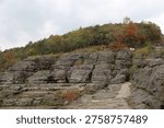 Layered, gray blocky stone structures form a rugged landscape. A stone pathway navigates the rock formations, leading toward a hilltop covered in autumn trees under an overcast sky.