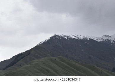 A layered mountain landscape rises under a heavy, overcast sky. In the foreground, a series of rolling, green-grey hillsides feature deep, undulating ridges and grassy slopes that tilt upward toward the left.

Behind these lower green slopes, a much larger, darker mountain mass dominates the upper background. Its craggy, rugged peaks are dusted with patches of white snow along the highest crests, contrasting against the dark stone. The flat, muted daylight casts subtle shadows throughout the valleys and ridges.