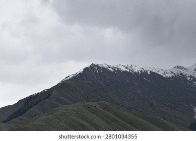 A layered mountain landscape rises under a heavy, overcast sky. In the foreground, a series of rolling, green-grey hillsides feature deep, undulating ridges and grassy slopes that tilt upward toward the left.

Behind these lower green slopes, a much larger, darker mountain mass dominates the upper background. Its craggy, rugged peaks are dusted with patches of white snow along the highest crests, contrasting against the dark stone. The flat, muted daylight casts subtle shadows throughout the valleys and ridges.