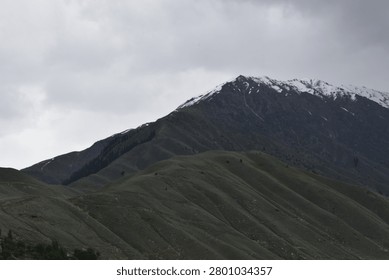 A layered mountain landscape rises under a heavy, overcast sky. In the foreground, a series of rolling, green-grey hillsides feature deep, undulating ridges and grassy slopes that tilt upward toward the left.

Behind these lower green slopes, a much larger, darker mountain mass dominates the upper background. Its craggy, rugged peaks are dusted with patches of white snow along the highest crests, contrasting against the dark stone. The flat, muted daylight casts subtle shadows throughout the valleys and ridges.