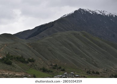 A layered mountain landscape rises under a heavy, overcast sky. In the foreground, a series of rolling, green-grey hillsides feature deep, undulating ridges and grassy slopes that tilt upward toward the left.

Behind these lower green slopes, a much larger, darker mountain mass dominates the upper background. Its craggy, rugged peaks are dusted with patches of white snow along the highest crests, contrasting against the dark stone. The flat, muted daylight casts subtle shadows throughout the valleys and ridges.