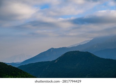 Layered mountain range under soft, cloudy sky. Forests cover rolling hillsides. Blue and green tones dominate landscape