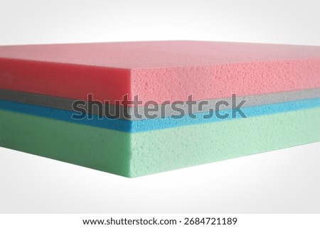 A layered sample of foam material showcasing distinct colors including pink, gray, light blue, and mint green. The texture of the foam is visible, with small air bubbles.