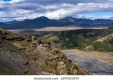 Layered volcanic cliffs drop to a braided ash gray river in Iceland. Snow dusted peaks near Thorsmork and Katla rise under midday light, with green moss and basalt.