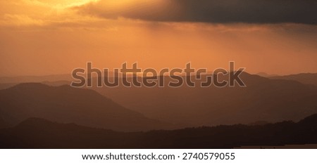 Layers of distant mountain silhouettes under a warm orange sky with the sun setting, creating a serene and vast natural landscape. Stormy dramatic sky, storm clouds, bright sunrays
