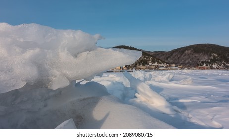 Layers of icy snow lie in disarray on a frozen lake. Close-up. In the distance, on the shore, you can see a wooded mountain range, a village. Blue sky. A sunny winter day. Baikal