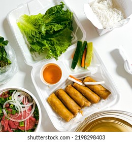 Layflat-style Photo Of Vietnamese Takeout Food, Including Deep Fried Pork Egg Rolls And Beef Pho Soup.