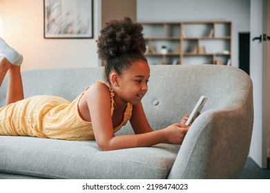 Laying Down On Sofa With Smartphone In Hands. Cute Black Girl In Casual Clothes Is At Home At Daytime.