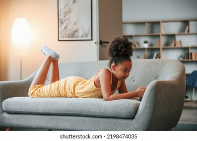 Laying Down On Sofa With Smartphone In Hands. Cute Black Girl In Casual Clothes Is At Home At Daytime.