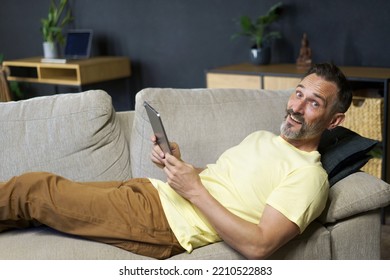 Laying On The Couch Middle Aged Freelancer Man Use Digital Tablet To Read Social Media Or Working Offline From Home Wearing Casual. Relaxed Freelancer Man Laying On Sofa Making Business Uses Gadget. 