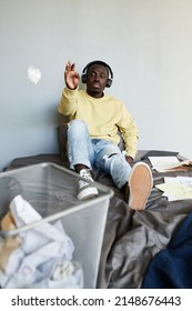 Lazy African-American Student Boy In Wireless Headphones Sitting On Bed And Throwing Crumpled Paper Into Trash Can While Wasting Time