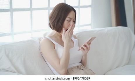 Lazy Asian Woman Face Yawning Using Mobile Smart Phone On Sofa Couch At Home. Female In White Living Room Yawn On Day Time Need Rest. Tired And Exhausted From Workload Sleep Fall Asleep And Nap.