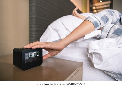 Lazy Asian Young Woman Trying To Snooze Awaking Digital Alarm Clock On The Table Beside The Bed Close Up, Woman Wake Up Late In Morning Up To 8.00 Am. Tried Woman Sleeping On The Bed In Bedroom.