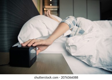 Lazy Asian Young Woman Trying To Snooze Awaking Digital Alarm Clock On The Table Beside The Bed Close Up, Woman Wake Up Late In Morning Up To 8.00 Am. Tried Woman Sleeping On The Bed In Bedroom.