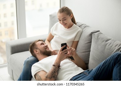 Lazy Bearded Man Lying On Lover Knees Showing Girlfriend Funny Video On Smartphone, Millennial Boyfriend Relaxing Playing Games On Phone, Couple Rest On Cozy Couch Having Fun Using Gadget