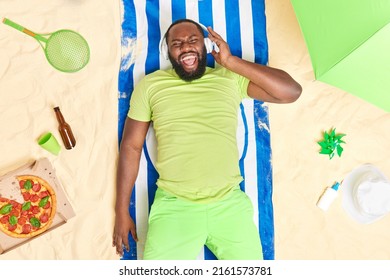Der hellbärtige junge Mann wirft am Sandstrand ein Handtuch auf, hört Musik über Kopfhörer und genießt Sommerferien Pizza trinkt kalte Biersonnenbäder an heißen Tagen am Meer. Perfekter Urlaub auf See