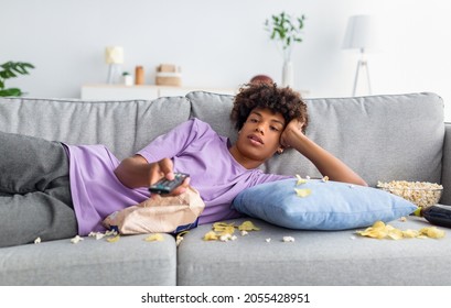 Lazy black teen guy lying on sofa with scattered food, holding TV controller, watching dull movie on television at home. Bored African American adolescent killing time indoors during covid lockdown - Shutterstock ID 2055428951