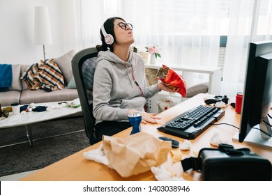 Lazy College Girl In Messy Dirty Bedroom With Order In Junk Food Snack Enjoy Unhealthy Life On Summer Break. Happy Young Asian Weird Woman With Headphones And Glasses Laughing Binge Watching Online