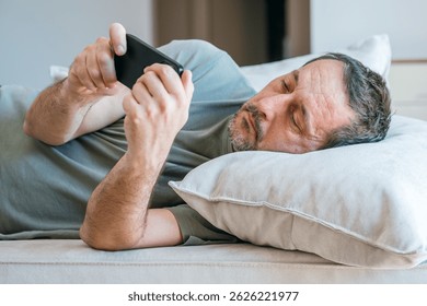 Lazy couch potato guy using mobile phone while laying on living room sofa, selective focus