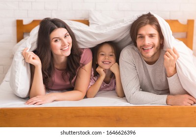 Lazy Family Weekend. Mom, Dad And Cute Little Daughter Hiding Under Blanket, Lying On Bed At Home And Looking At Camera