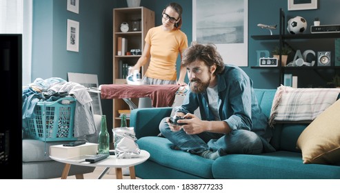 Lazy Husband Playing Videogames And Relaxing On The Sofa While His Wife Is Doing Household Chores, Gender Inequality Concept