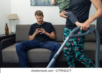 Lazy Husband Sitting In Couch And Using Phone And Not Helping Wife While She Cleaning House With Child On Her Lap