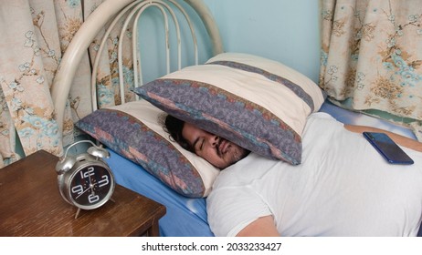 A lazy and jobless guy in his 30s covers his face with a pillow, and his phone on top of his stomach. Unemployed bum not getting up of bed.