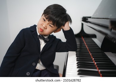 Lazy little boy in black suit sitting at grand piano makes funny face. Cute boring kid pianist feels tired to practice playing classic keyboard. Playful child musician refuses to perform a concert.