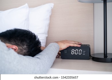 Lazy Male Office Worker Sleeping On Bed In Bedroom, Stopping Alarm Clock In The Morning On Weekday. Man Put Hand On Digital Clock Button To Silent The Voice And Feeling Bore To Wake Up, Feels Sleepy.