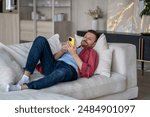 Lazy man lies on sofa in living room, uses phone, thoughtlessly meaninglessly scrolling through feed of social networks, absorbing information, content. Tired calm guy resting on couch with smartphone