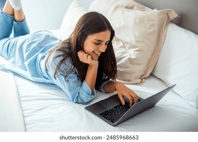 Lazy modern young woman using social media on laptop laying on bed in morning wake up wearing casual clothes and smiling at the display. Working remote with computer and wireless connection in hotel  - Powered by Shutterstock