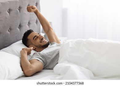Lazy Morning. Happy Pleased Middle Eastern Man Stretching In Bed After Nice Sleep, Cheerful Young Arab Guy Waking Up With Good Mood, Relaxing In Light Bedroom, Enjoying Weekend Pastime, Copy Space - Shutterstock ID 2104377368